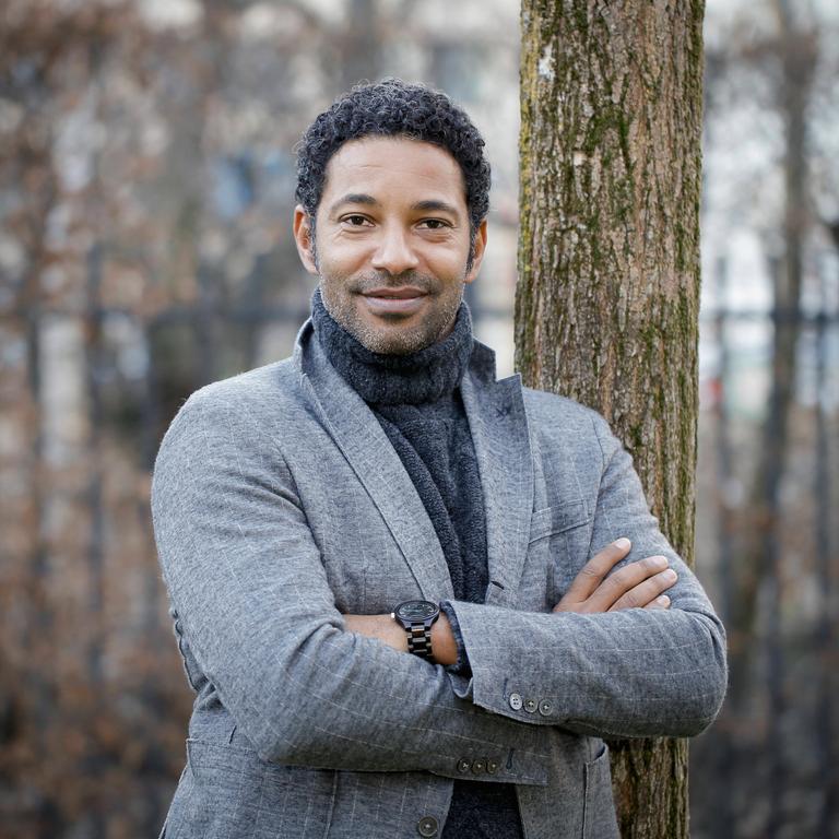 Der Schauspieler und Produzent Tyron Ricketts steht mit einem wollenen Jackett und grauem Rollkragenpullover bekleidet in einem Garten und blickt in die Kamera.