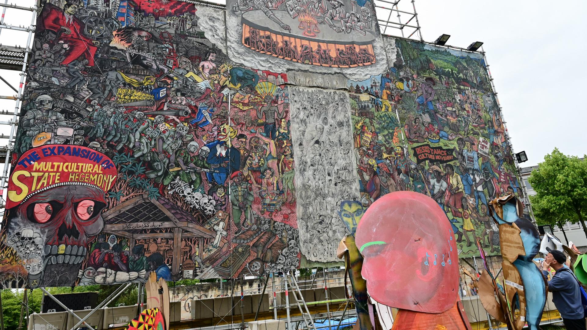 Auf dem etwa 10 x 10 Meter großen Bild der indonesischen Künstlergruppe Taring Padi ist ein Gewimmel verschiedenster Figuren zu sehen, links unten ein Totenkopf und auf dessen Kopfbedeckung die Aufschrift "The Expansion of Multicultural State Hegemony". Auf dem etwa 10 x 10 Meter großen Bild der indonesischen Künstlergruppe Taring Padi ist ein Gewimmel verschiedenster Figuren zu sehen, links unten ein Totenkopf und auf dessen Kopfbedeckung die Aufschrift "The Expansion of Multicultural State Hegemony".