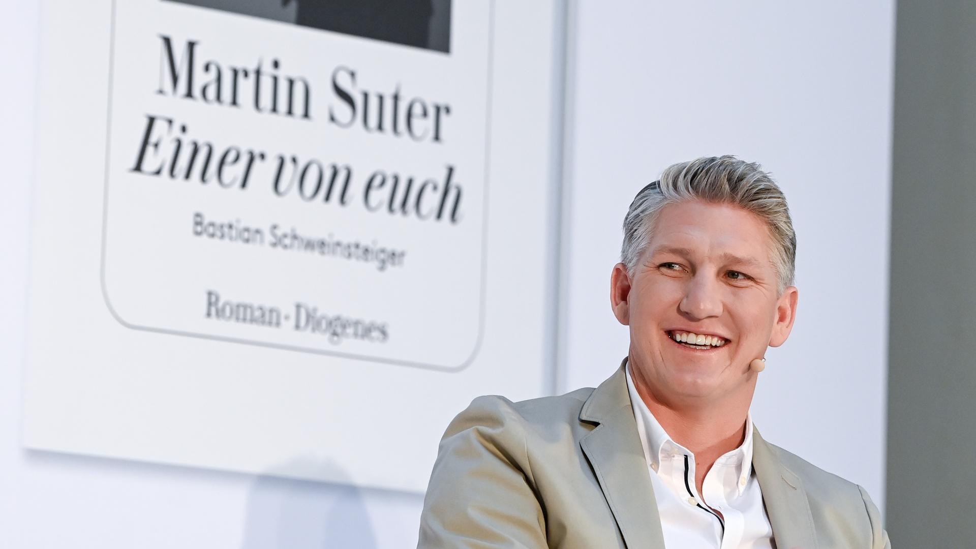 Der Fußballer Bastian Schweinsteiger sitzt in einem hellen Sakko auf einem Podium, hinter ihm an der Wand: der Buchumschlag des biografischen Romans "Einer von uns", in dem der Bestsellerautor Martin Suter aus Schweinsteigers Leben erzählt. Der Fußballer Bastian Schweinsteiger sitzt in einem hellen Sakko auf einem Podium, hinter ihm an der Wand: der Buchumschlag des biografischen Romans "Einer von uns", in dem der Bestsellerautor Martin Suter aus Schweinsteigers Leben erzählt.