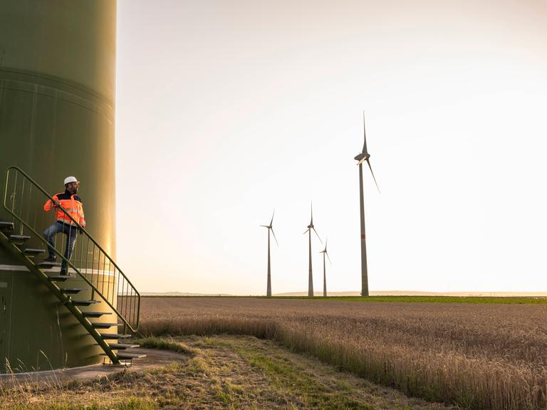 Eine Windkraftanlage. Ein Bauarbeiter steht auf einer Treppe zur Anlage, daneben Windräder auf einem Feld.