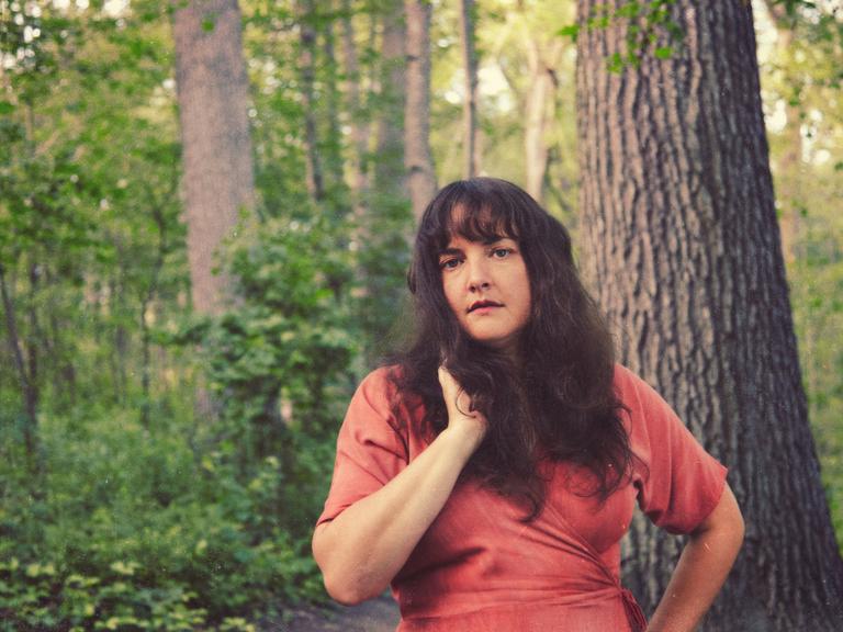 Sängerin Abigail Lapell steht in einem frischen, grünen Wald vor einem großen Baum in rotem Wickelkleid und offenen dunklen, leicht welligen Haaren.