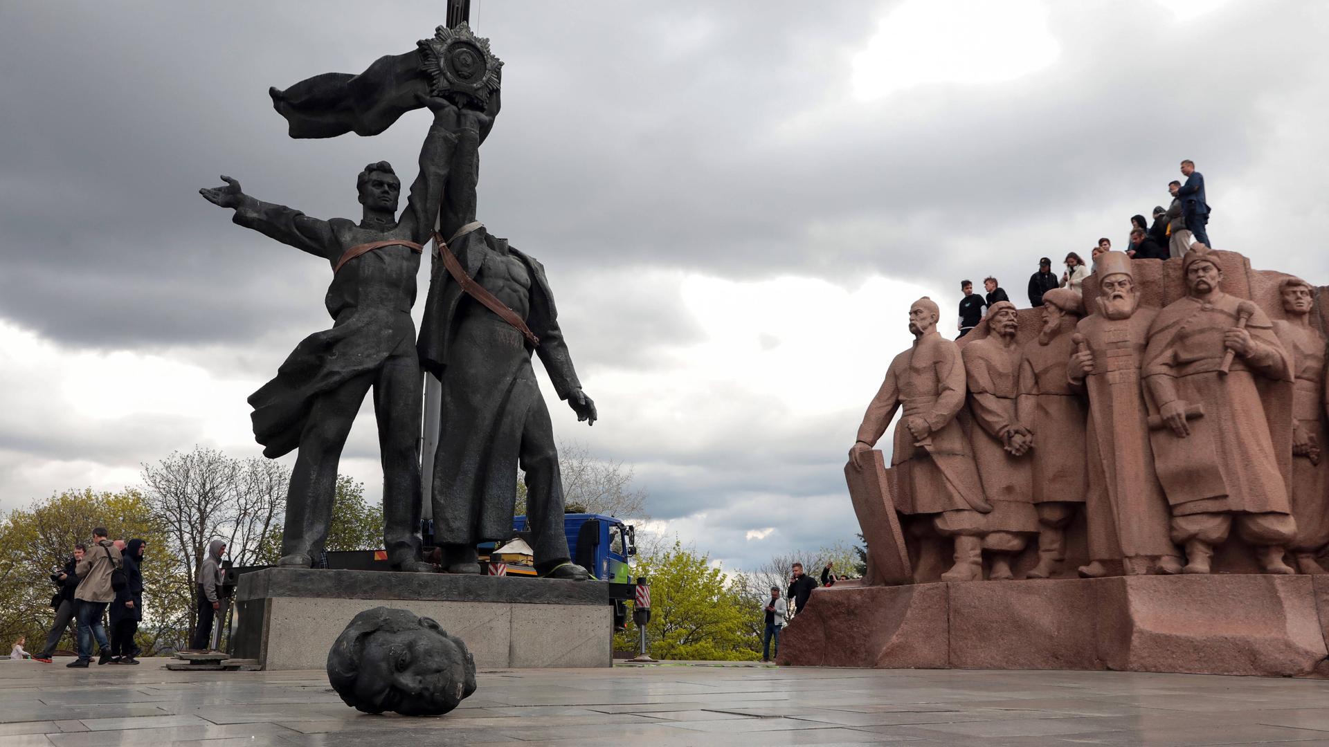 Zwei schwarze Männerstatuen stehen nebeneinander und halten in der Mitte eine Fahne fest. Im Vordergrund liegt der Kopf der rechten Statue. Rechts sind Heldengestalten aus rötlichem Stein zu sehen.