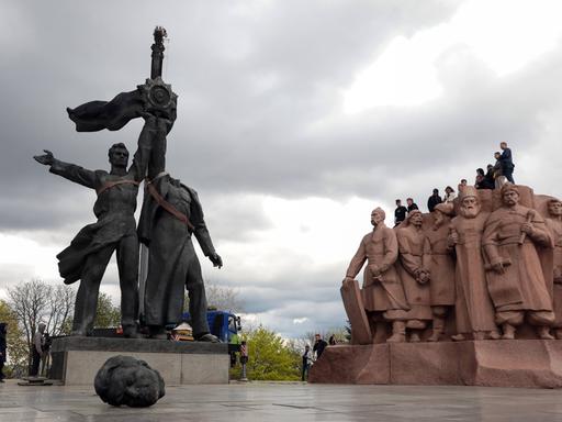 Zwei schwarze Männerstatuen stehen nebeneinander und halten in der Mitte eine Fahne fest. Im Vordergrund liegt der Kopf der rechten Statue. Rechts sind Heldengestalten aus rötlichem Stein zu sehen.