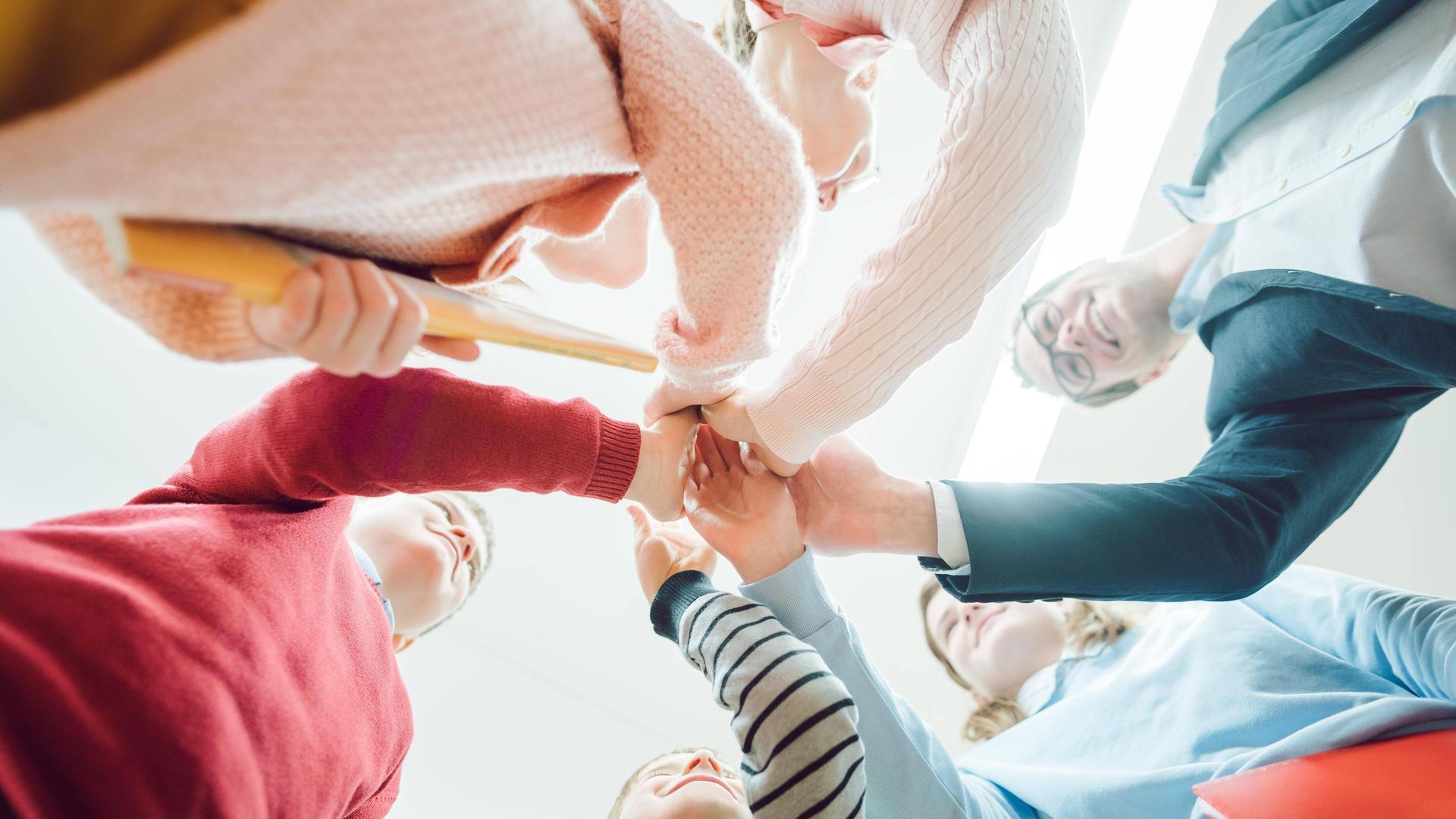 Schülerinnen und Schüler bilden mit ihrer Lehrerin einen Kreis und legen die Hände aufeinander, mit Perspektive von unten. Schülerinnen und Schüler bilden mit ihrer Lehrerin einen Kreis und legen die Hände aufeinander, mit Perspektive von unten.