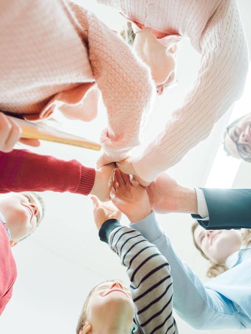Schülerinnen und Schüler bilden mit ihrer Lehrerin einen Kreis und legen die Hände aufeinander, mit Perspektive von unten.