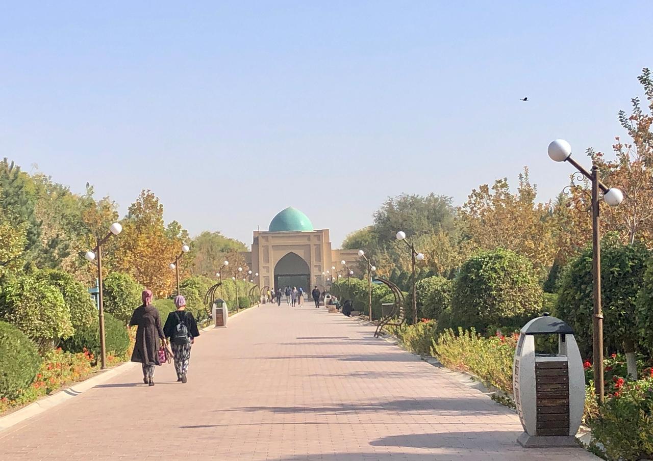 Zwei Frauen gehen einen gepflasterten Weg entlang an dessen Ende eine Moschee steht mit grünem Kuppeldach. Die Sonne scheint.