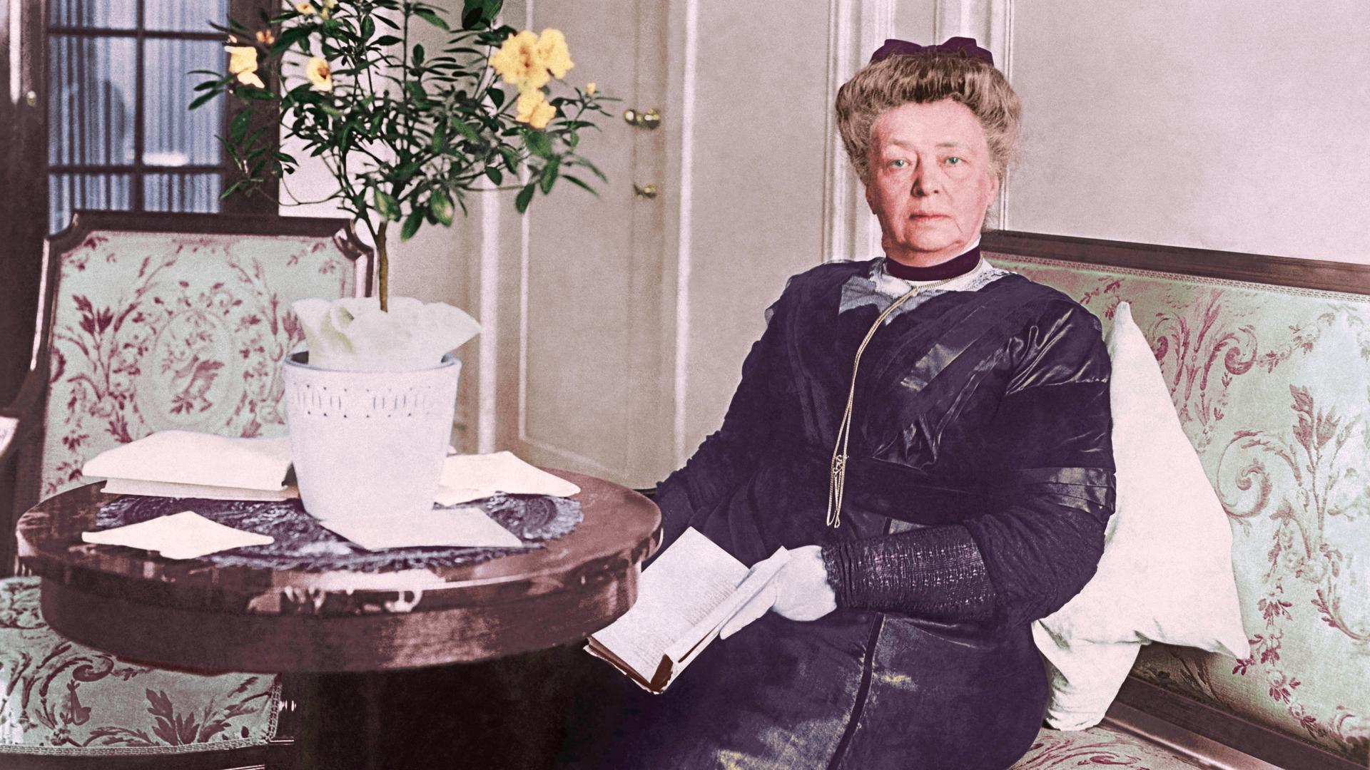 Bertha von Suttner sitzt in der Kulisse eines bürgerlichen Haushalts um 1900 in formaler Kleidung auf einem Sofa mit Blumenmuster an einem runden Tisch.