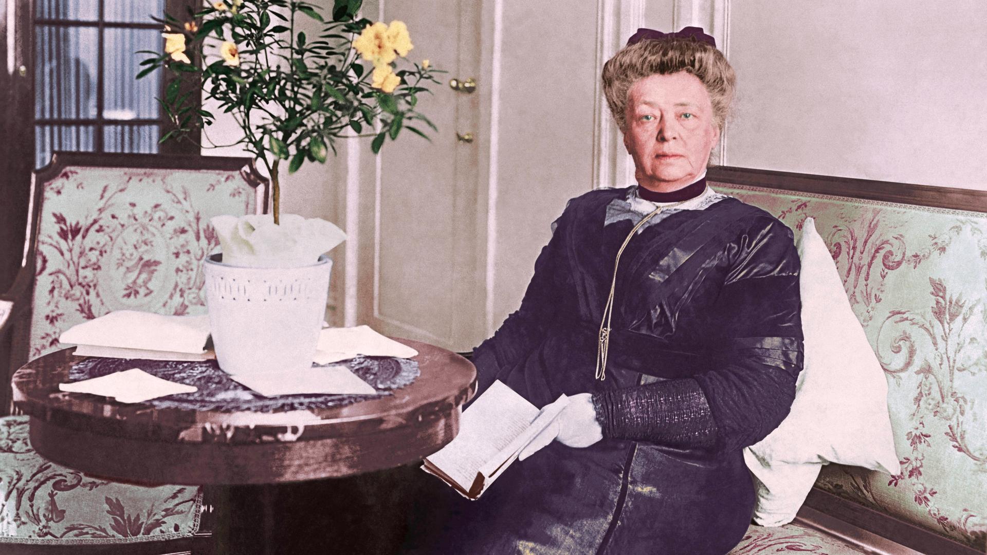 Bertha von Suttner sitzt in der Kulisse eines bürgerlichen Haushalts um 1900 in formaler Kleidung auf einem Sofa mit Blumenmuster an einem runden Tisch. Bertha von Suttner sitzt in der Kulisse eines bürgerlichen Haushalts um 1900 in formaler Kleidung auf einem Sofa mit Blumenmuster an einem runden Tisch.