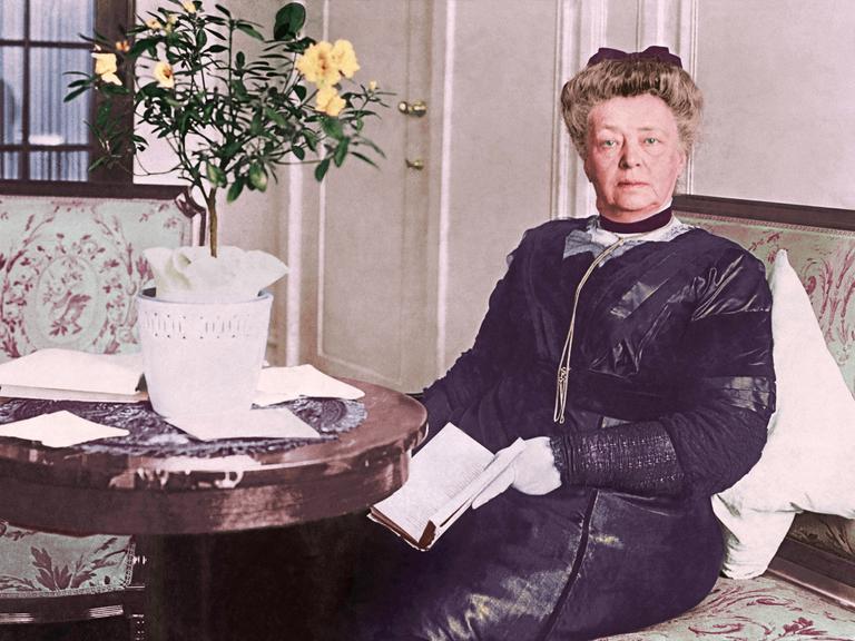 Bertha von Suttner sitzt in der Kulisse eines bürgerlichen Haushalts um 1900 in formaler Kleidung auf einem Sofa mit Blumenmuster an einem runden Tisch.