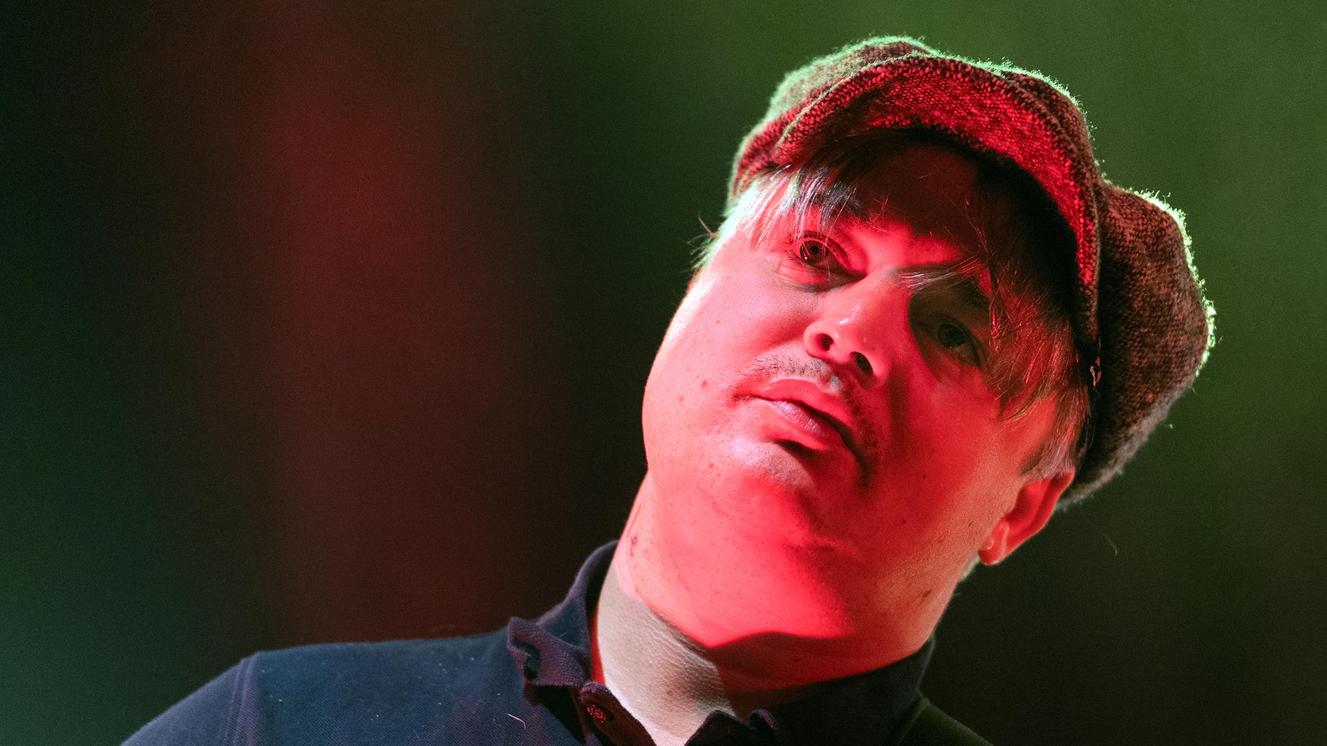 Pete Doherty mit einer Schiebermütze auf der Bühne. Sein Gesicht wird von einem Bühnenscheinwerfer rötlich angestahlt. Pete Doherty mit einer Schiebermütze auf der Bühne. Sein Gesicht wird von einem Bühnenscheinwerfer rötlich angestahlt.