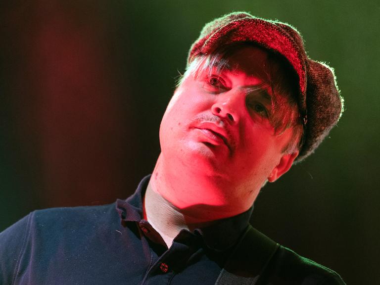 Pete Doherty mit einer Schiebermütze auf der Bühne. Sein Gesicht wird von einem Bühnenscheinwerfer rötlich angestahlt.