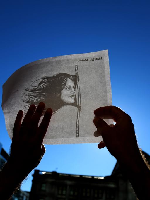 Eine Person hält eine weiße Fahne in die Luft auf der eine Zeichnung von Mahsa Amini zu sehen ist.