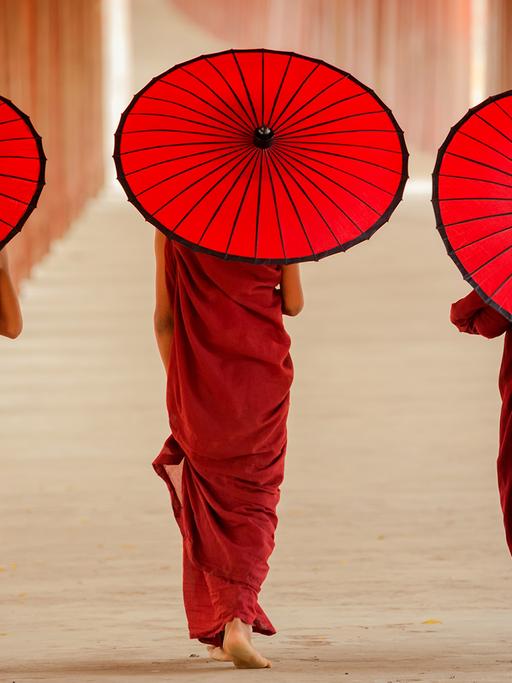 Drei Zen-Mönche mit je einem roten Sonnenschirm gehen mit dem Rücken zur Kamera einen Klostergang entlang. 