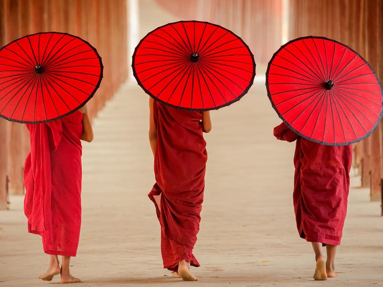 Drei Zen-Mönche mit je einem roten Sonnenschirm gehen mit dem Rücken zur Kamera einen Klostergang entlang. 