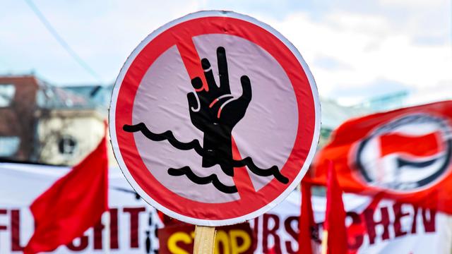 Ein rundes Schild mit der einem Piktogramm einer rot durchgestrichenen Hand in den Wellen versinkend, ist auf einer Demonstration in Stuttgart zu sehen.