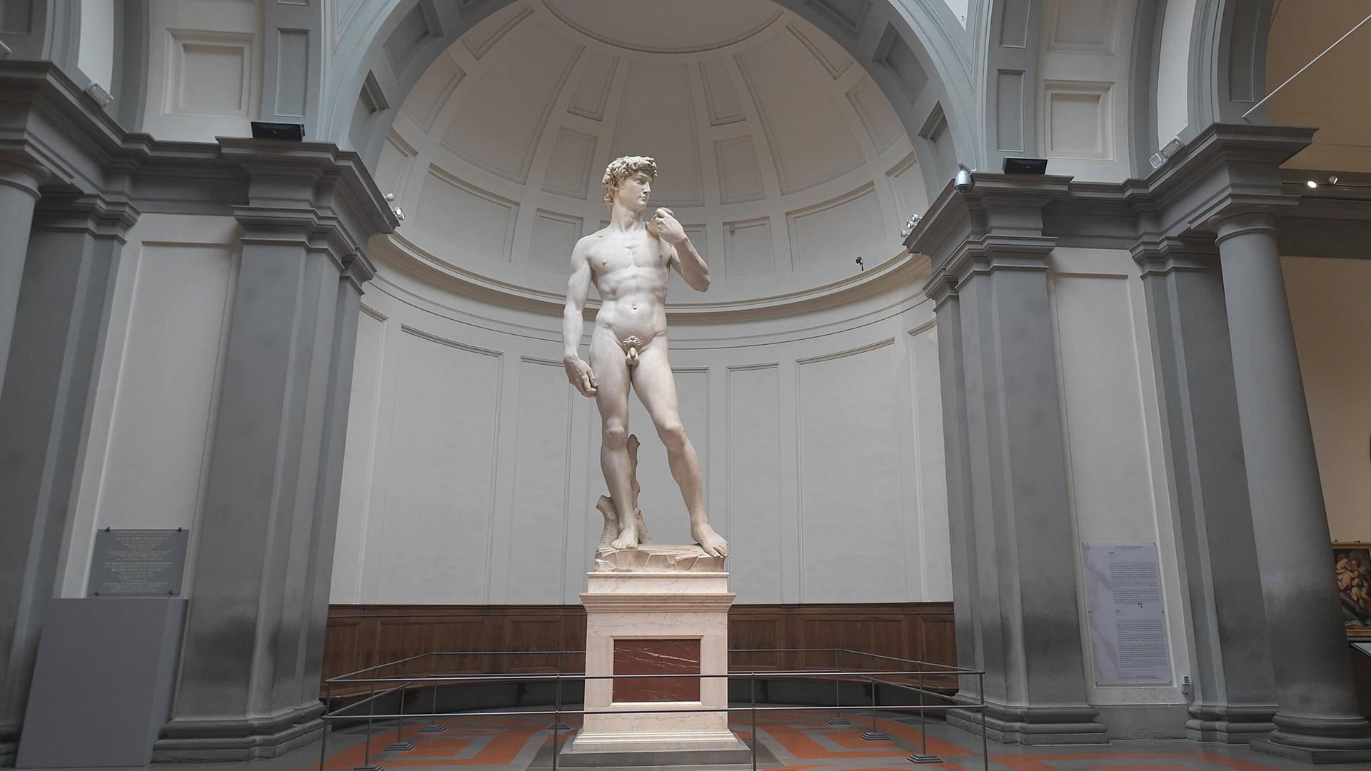 Die übergroße weiße Skulptur eines nackten Mannes steht in entspannter Pose auf einem Sockel allein in einem Museumsraum. Die übergroße weiße Skulptur eines nackten Mannes steht in entspannter Pose auf einem Sockel allein in einem Museumsraum.