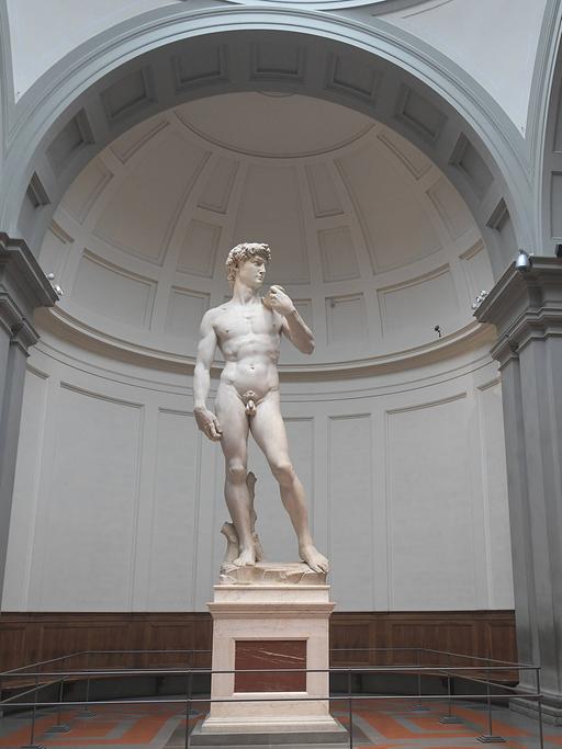 Die übergroße weiße Skulptur eines nackten Mannes steht in entspannter Pose auf einem Sockel allein in einem Museumsraum.