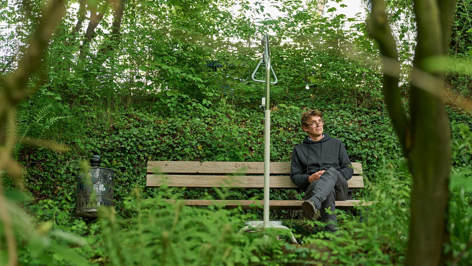 Ein Mann sitzt auf einer Bank in einem Park. Neben ihm steht ein graues Gestell, mit dem man sich im Krankenbett hochziehen kann. Er lauscht einer Klanginstallation. Ein Mann sitzt auf einer Bank in einem Park. Neben ihm steht ein graues Gestell, mit dem man sich im Krankenbett hochziehen kann. Er lauscht einer Klanginstallation.
