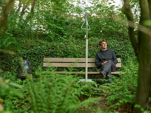 Ein Mann sitzt auf einer Bank in einem Park. Neben ihm steht ein graues Gestell, mit dem man sich im Krankenbett hochziehen kann. Er lauscht einer Klanginstallation.