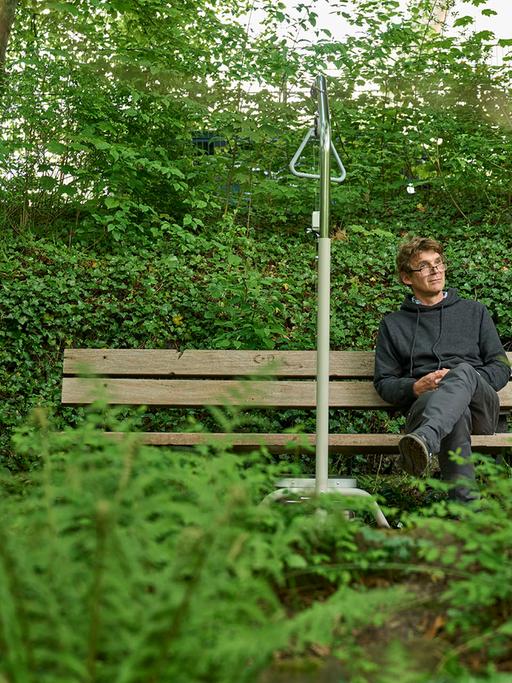 Ein Mann sitzt auf einer Bank in einem Park. Neben ihm steht ein graues Gestell, mit dem man sich im Krankenbett hochziehen kann. Er lauscht einer Klanginstallation.