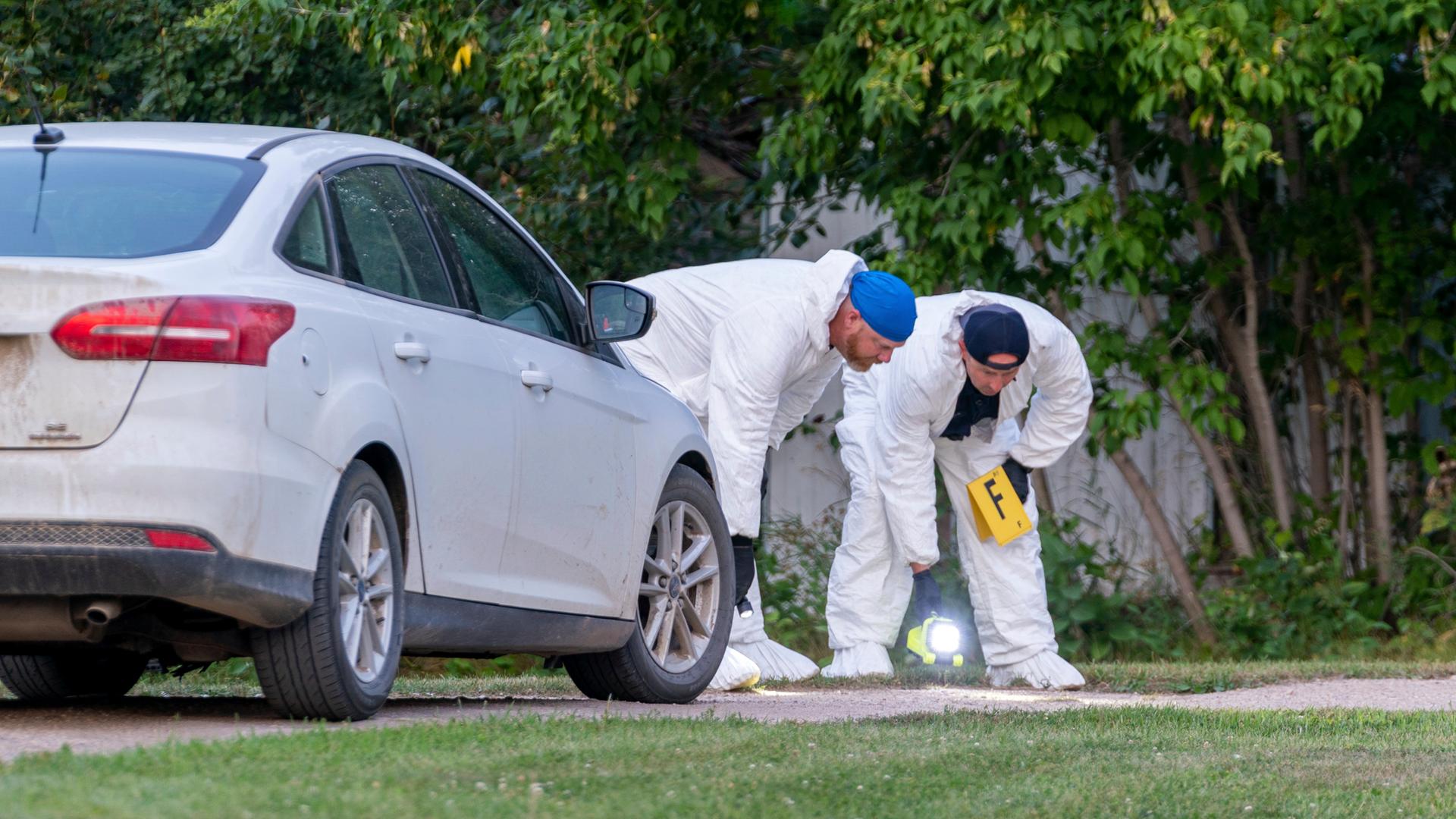 Zwei Menschen in weißen Anzügen beugen sich zum Boden. Daneben steht ein weißes Auto. Zwei Menschen in weißen Anzügen beugen sich zum Boden. Daneben steht ein weißes Auto.