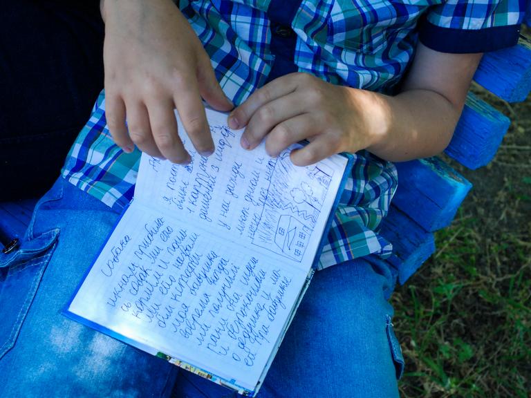 Der achtjährige Egor hält ein kleines blaues Buch in seiner Hand. Er schrieb in Mariupol Tagebuch, als um die Stadt blutig gekämpft wurde. Auf der aufgeschlagenen Seite steht neben einer kleinen Kinderzeichnung: "Ich habe gut geschlafen, habe gelächelt und 25 Seiten gelesen. Außerdem ist mein Großvater gestorben am 26.April. Saporischschja, 3. Juni 2022."