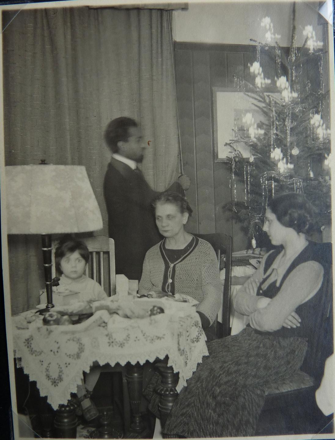 Eine Frau sitzt mit einem etwa vierjährigen Jungen und einem etwa 14-jährigen Mädchen um einen Tisch. Im Hintergrund zündet ein Mann Kerzen an einem Weihnachtsbaum an.