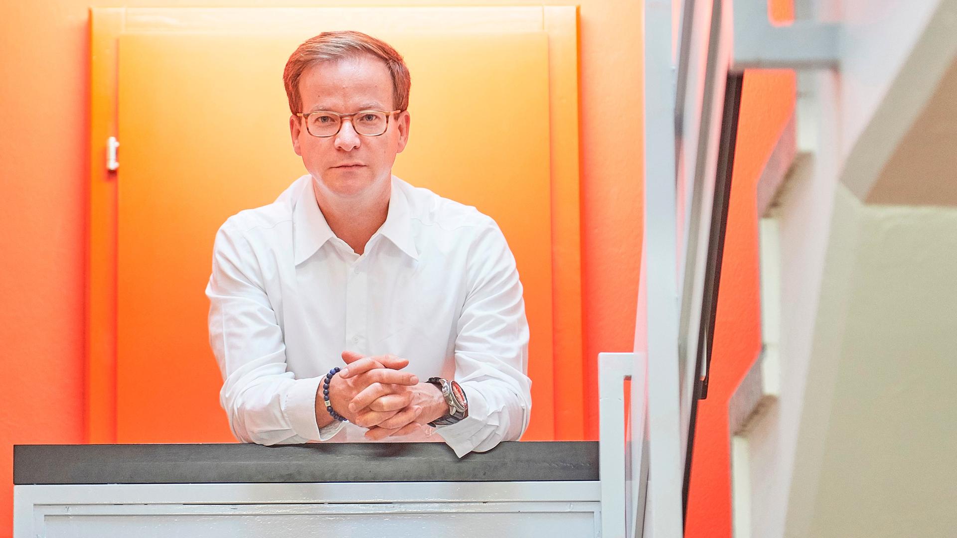 Ein schlanker, weißer Mann mit dunkelblonden Haaren und Brille posiert fürs Foto in einem Treppenhaus, die Wand und Tür hinter ihm ist leuchtend orange. Der Mann trägt ein weißes Hemd, ein Armband und eine auffällige Armbanduhr. Ein schlanker, weißer Mann mit dunkelblonden Haaren und Brille posiert fürs Foto in einem Treppenhaus, die Wand und Tür hinter ihm ist leuchtend orange. Der Mann trägt ein weißes Hemd, ein Armband und eine auffällige Armbanduhr.
