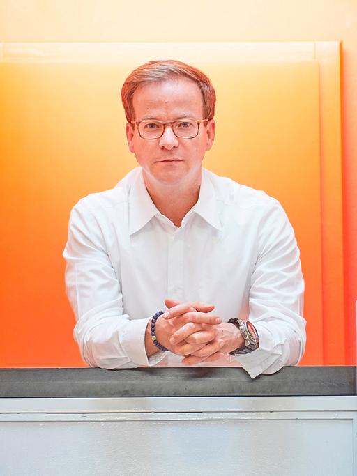 Ein schlanker, weißer Mann mit dunkelblonden Haaren und Brille posiert fürs Foto in einem Treppenhaus, die Wand und Tür hinter ihm ist leuchtend orange. Der Mann trägt ein weißes Hemd, ein Armband und eine auffällige Armbanduhr.