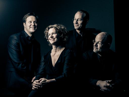 Die vier Musiker, eine Frau umringt von drei Männern, lächlel versonnen, während sie eng in dunkler Kleidung beieinander sitzen.