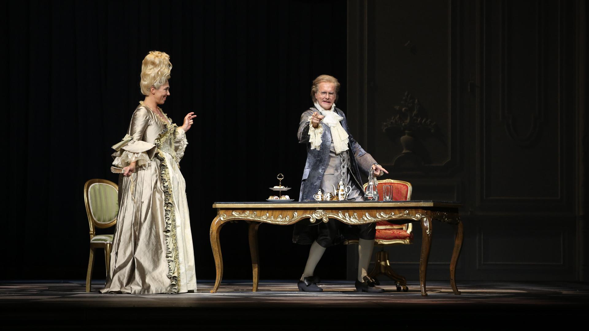 Harald Schmidt steht als König Ludwig XV. hinter einem Schreibtisch und zeigt ins Publikum. Und Annette Dasch als Gräfin Dubarry steht neben dem Tisch und schaut ihn an.