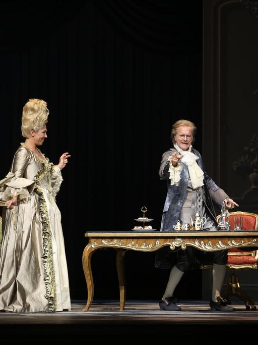 Harald Schmidt steht als König Ludwig XV. hinter einem Schreibtisch und zeigt ins Publikum. Und Annette Dasch als Gräfin Dubarry steht neben dem Tisch und schaut ihn an.