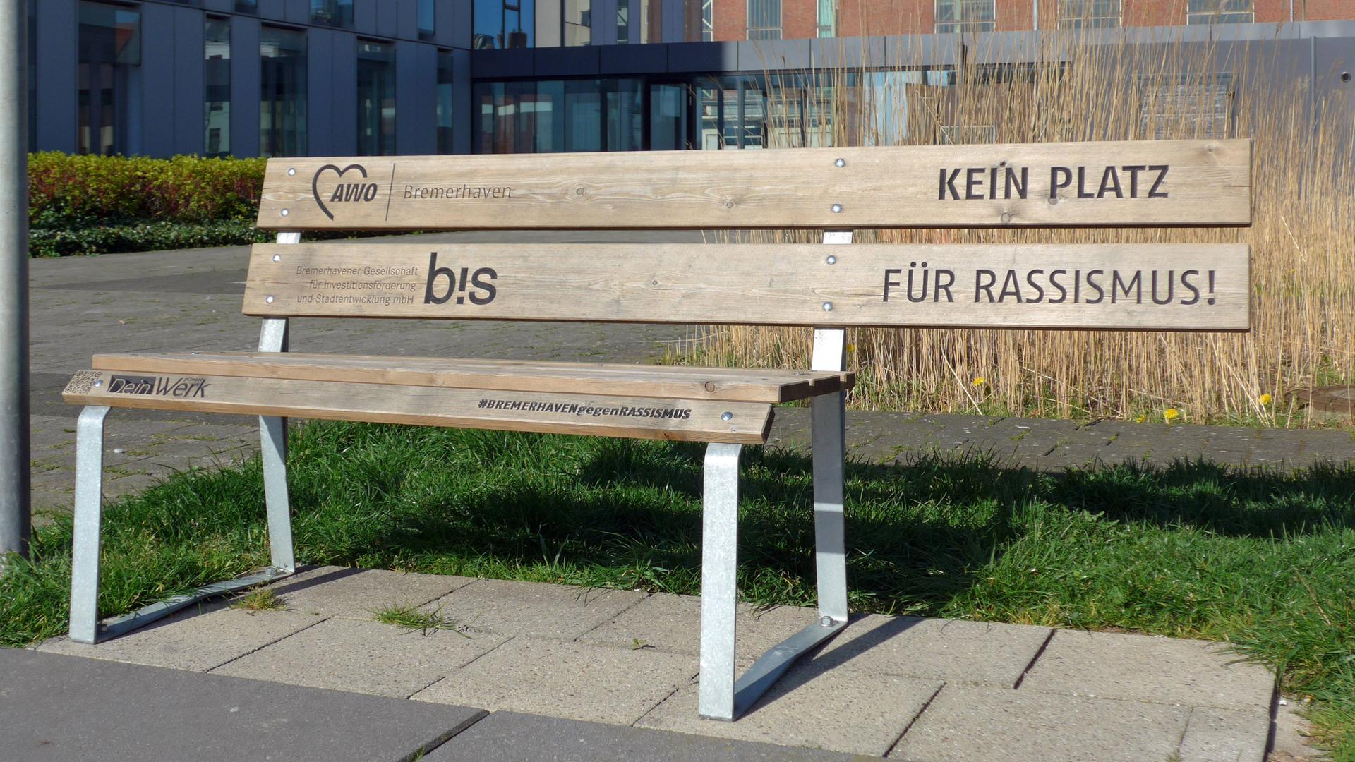 Die Rückenlehne einer Holzbank ragt über die Sitzfläche hinaus. Dort, wo niemand sitzen kann, steht auf der Lehne: "Kein Platz für Rassismus". Die Rückenlehne einer Holzbank ragt über die Sitzfläche hinaus. Dort, wo niemand sitzen kann, steht auf der Lehne: "Kein Platz für Rassismus".