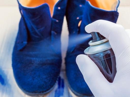 Eine Person sprüht, Gumminhandschuhe tragend, blaue Wildlederschuhe mit Imprägnierspray ein.