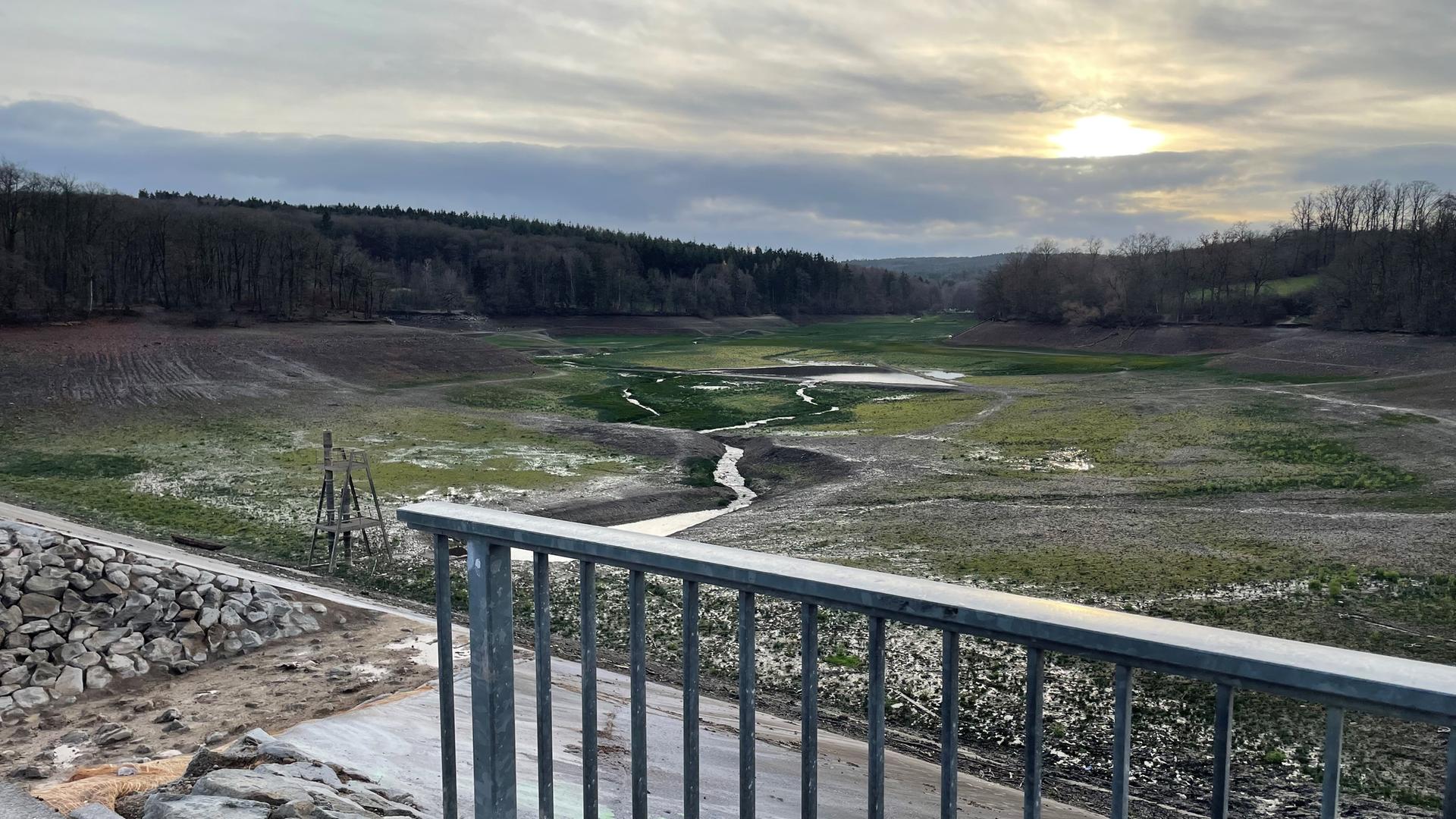 Blick vom Staudamm aus in die leere Steinbachtalsperre. Darin sieht man einen kleinen Wasserlauf und Pfützen. Im Vordergrund steht ein Geländer. Blick vom Staudamm aus in die leere Steinbachtalsperre. Darin sieht man einen kleinen Wasserlauf und Pfützen. Im Vordergrund steht ein Geländer.