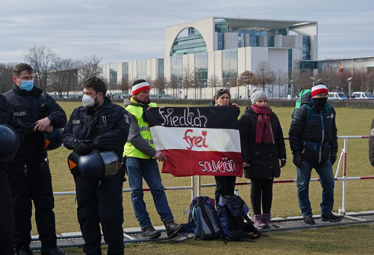 Vier Personen vor dem Reichstag in Berlin. Zwei halten ein schwarz-weiss-rotes Banner auf dem steht "friedlich, frei, souverän". Im Hintergrund ist das Bundeskanzleramt zu sehen.