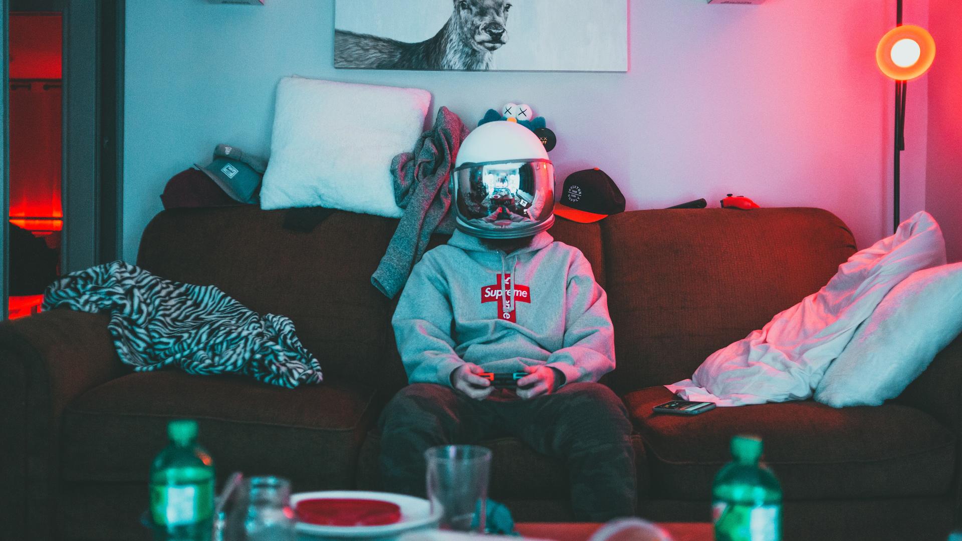Fotografie eines jungen Mannes auf der Couch, mit einem Helm, dessen verspiegeltes Visier geschlossen ist. Im Bildvordegrund sind Getränkeflaschen und Geschirr zu sehen, über der Couch hängt das Bild eines Rentiers mit Geweih. Fotografie eines jungen Mannes auf der Couch, mit einem Helm, dessen verspiegeltes Visier geschlossen ist. Im Bildvordegrund sind Getränkeflaschen und Geschirr zu sehen, über der Couch hängt das Bild eines Rentiers mit Geweih.