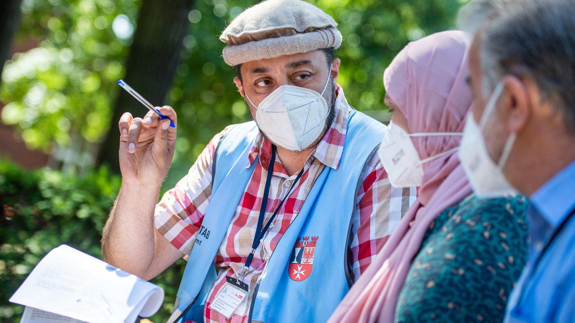 Taman Noor hält Papiere und einen Stift in der Hand. Er trägt eine hellblaue Weste mit dem Wappen des Berliner Bezirks Neukölln und spricht mit einer Frau mit Kopftuch und einem Mann. Alle tragen Masken. Taman Noor hält Papiere und einen Stift in der Hand. Er trägt eine hellblaue Weste mit dem Wappen des Berliner Bezirks Neukölln und spricht mit einer Frau mit Kopftuch und einem Mann. Alle tragen Masken.