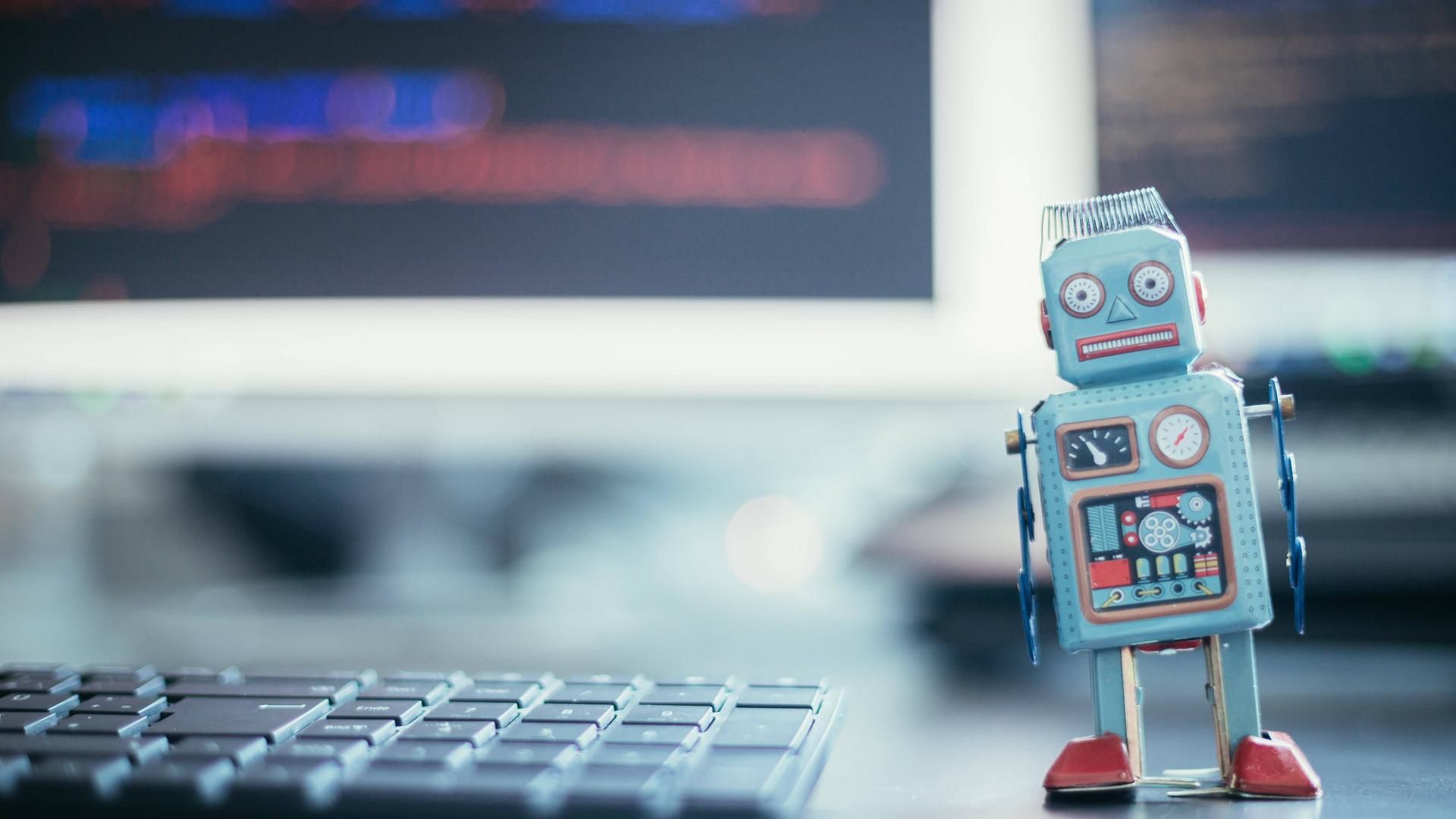 Symbolfoto: Ein kleiner Roboter steht neben einer Tastatur. Im Hintergrund unscharfe Computerbildschirme mit Texten. Symbolfoto: Ein kleiner Roboter steht neben einer Tastatur. Im Hintergrund unscharfe Computerbildschirme mit Texten.