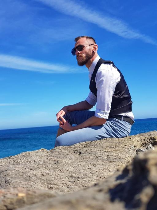 Ein junger Mann mit Sonnenbrille sitzt auf einem Felsen am Meer.