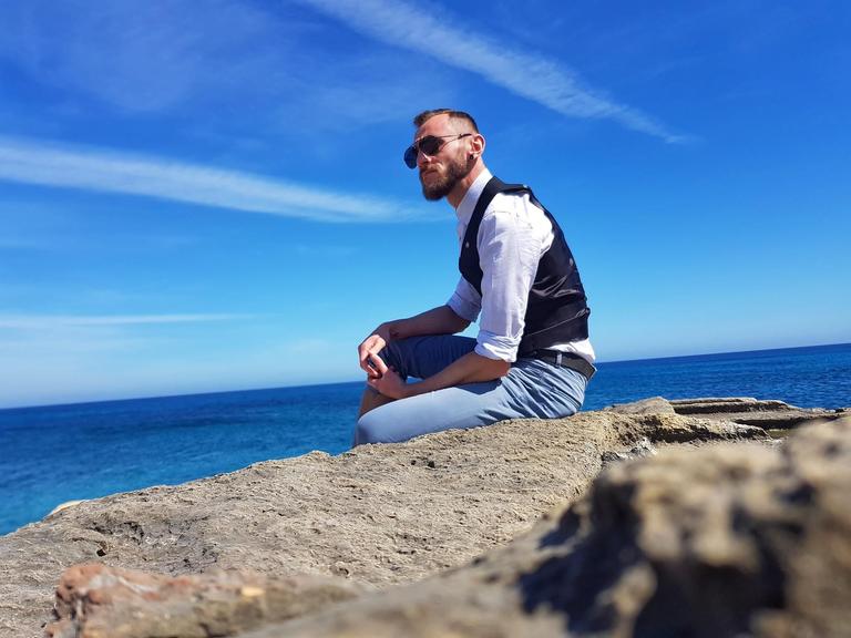 Ein junger Mann mit Sonnenbrille sitzt auf einem Felsen am Meer.