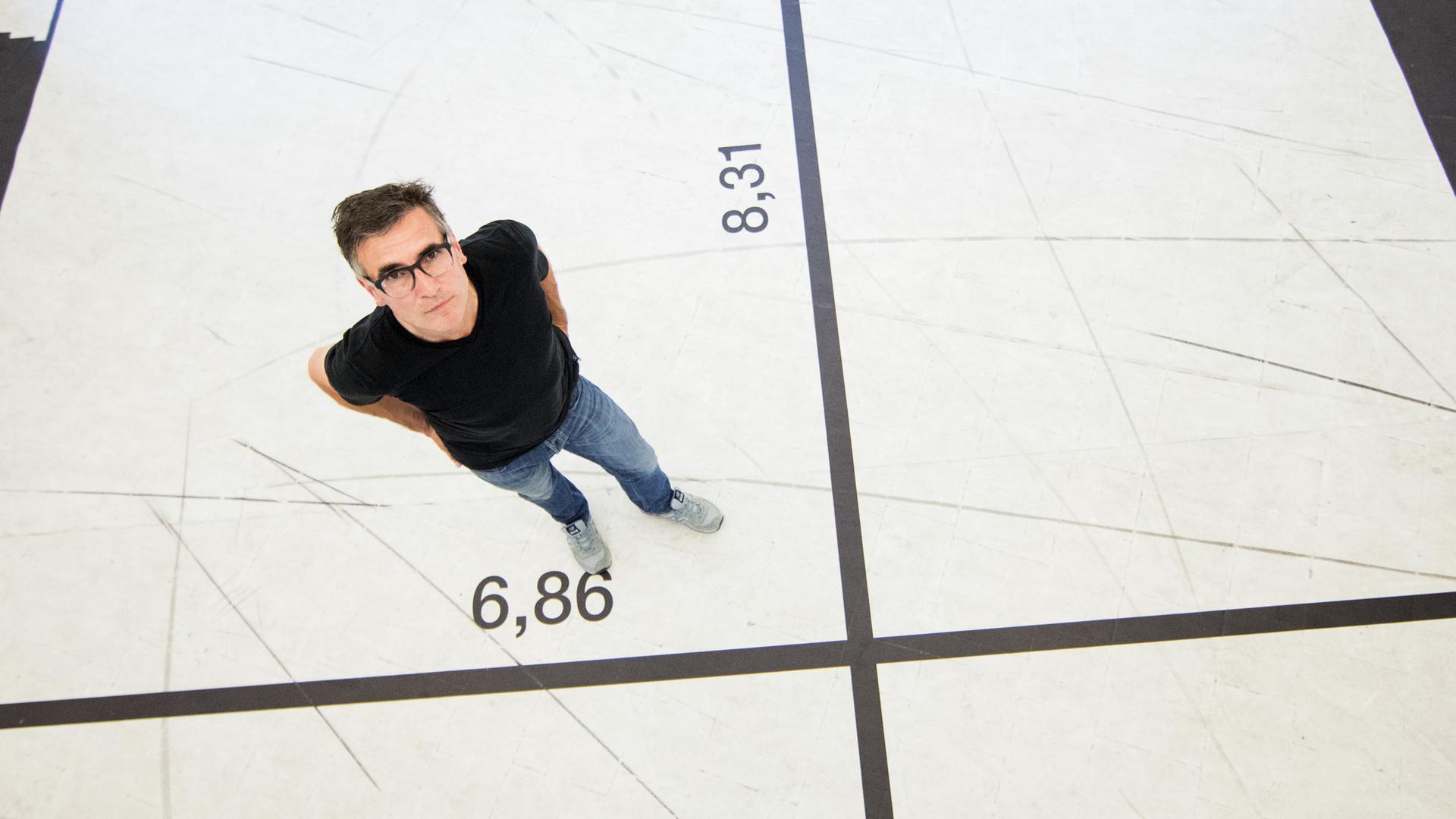 Ein Mann steht - von oben fotografiert - auf einem weißen Boden, der mit schwarzen Linien gekreuzt ist und an den Linien ist jeweils eine Dezimalzahl gedruckt.