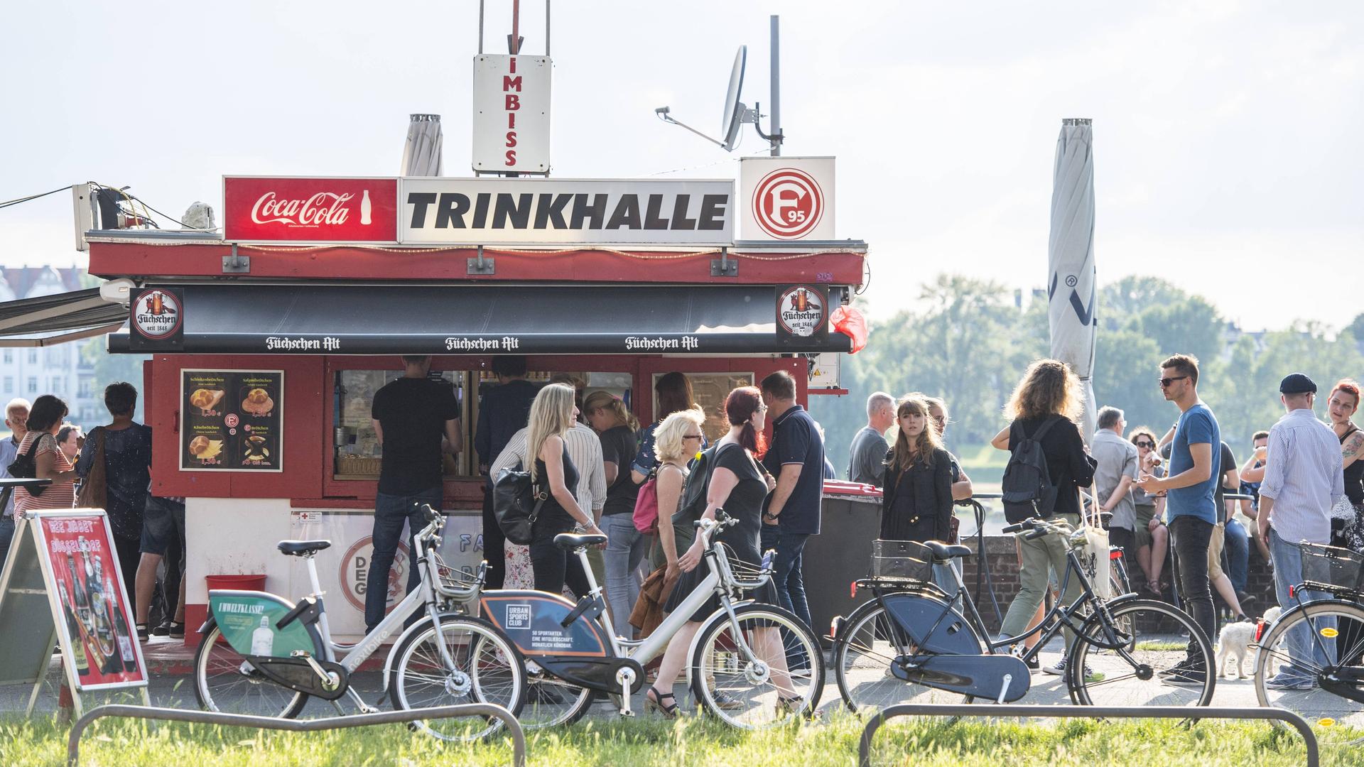 Ein Büdchen am Rheinufer in Düsseldorf, die Menschen stehen Schlange für ein Getränk an einem Sommerabend."Trinkhalle" und "Imbiss" steht auf dem Dach. Ein Büdchen am Rheinufer in Düsseldorf, die Menschen stehen Schlange für ein Getränk an einem Sommerabend."Trinkhalle" und "Imbiss" steht auf dem Dach.