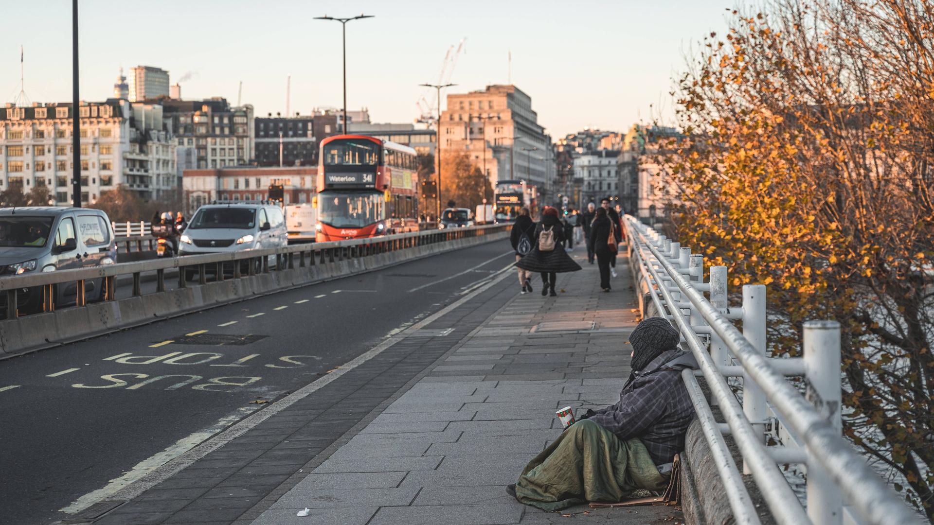 Großbritannien, London: Ein Bettler sitzt auf einer Brücke mit einem Becher für Kleingeld. Er ist dick eingepackt mit Mütze und Decke, im Hintergrund sind Passanten und der fließende Verkehr zu sehen.