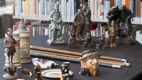 Auf einem Schreibtisch stehen verschiedene Objekte, die unter anderem menschliche Figuren in antisemitisch-klischeehafter Weise darstellen. 