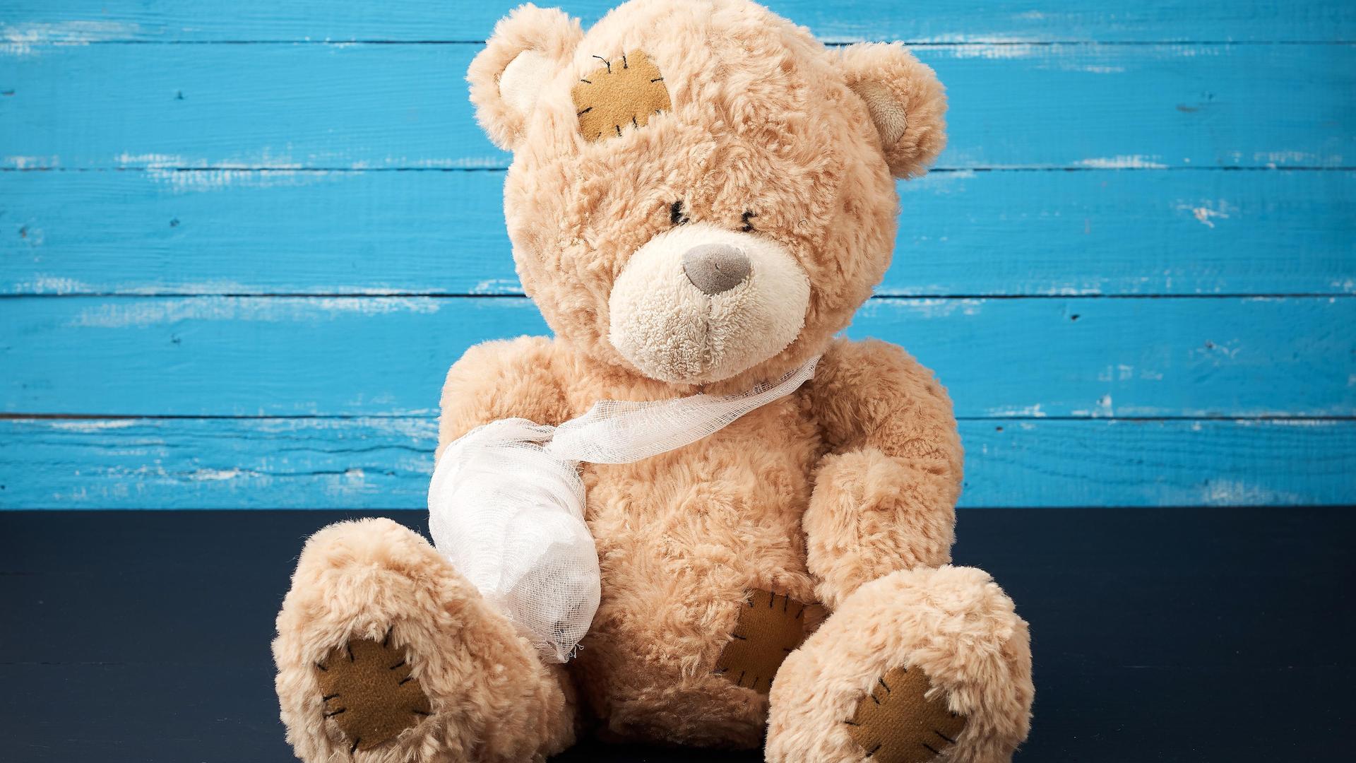 Ein brauner Teddy sitzt vor einer blauen Wand, seiner einer Arm hängt in einer Schlinge und ist eingegipst. Ein brauner Teddy sitzt vor einer blauen Wand, seiner einer Arm hängt in einer Schlinge und ist eingegipst.