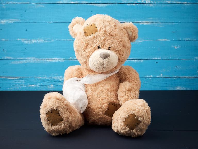 Ein brauner Teddy sitzt vor einer blauen Wand, seiner einer Arm hängt in einer Schlinge und ist eingegipst.