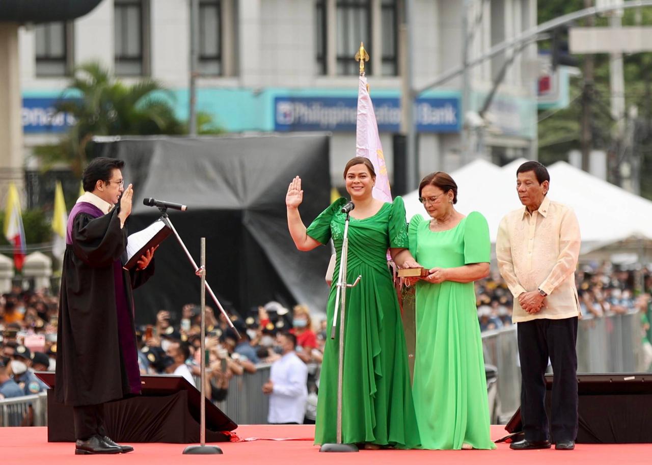 Zwei Frauen in grünen Kleidern stehen auf einer Bühne. Eine wird von einem Herrn in schwarzer Kleidung vereidigt, hebt die Rechte Hand zum Schwurd.