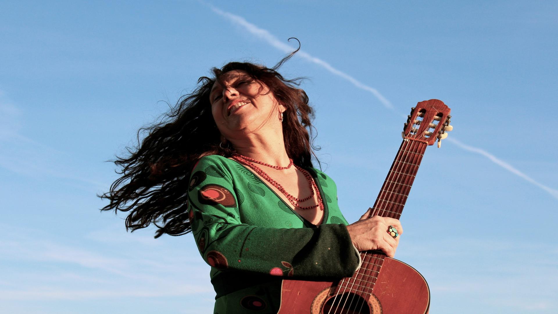 Zélia Fonseca dreht sich schwungvoll mit ihrer Gitarre und offenem Haar vor blauem Himmel. Zélia Fonseca dreht sich schwungvoll mit ihrer Gitarre und offenem Haar vor blauem Himmel.