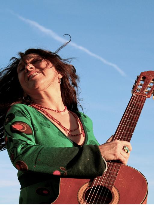 Zélia Fonseca dreht sich schwungvoll mit ihrer Gitarre und offenem Haar vor blauem Himmel. 